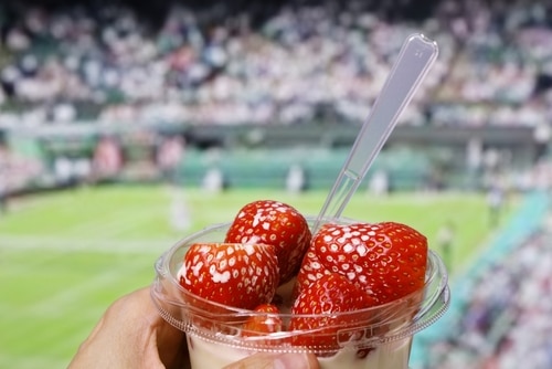 Erdbeeren mit Sahne im Vordergrund und der unscharfe Tennisplatz im Hintergrund.